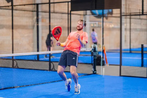 Riga, Letonya - 5 Nisan 2025 - Parlak pembe tişörtlü Padel oyuncusu, kapalı mavi bir kortta yapılan bir turnuva sırasında bir şutu kaçırdıktan ya da tamamladıktan sonra neşeli bir şekilde tepki verdi.