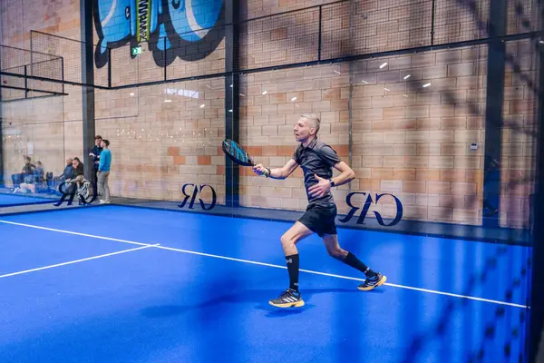 Riga, Letonya - 5 Nisan 2025 - Siyah spor giysili bir adam, arka planda cam duvarlar ve izleyiciler olan mavi bir iç sahada düzenlenen bir padel turnuvasında tekrar atış yapmak için ileri atıldı..