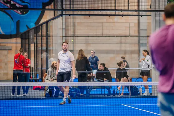Riga, Letonya - 5 Nisan 2025 - Padel turnuva sahnesi mavi sahada yürüyen bir erkek oyuncunun elinde raket, seyirciler ve katılımcılar camların arkasında görünür bir mekanda.