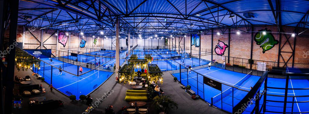 Riga, Latvia - April 5, 2025 - Panoramic view of a vibrant indoor padel tournament facility with multiple blue courts, players in action, and a central seating lounge under warm lighting.