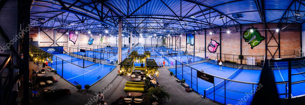 Riga, Latvia - April 5, 2025 - Panoramic view of a vibrant indoor padel tournament facility with multiple blue courts, players in action, and a central seating lounge under warm lighting.