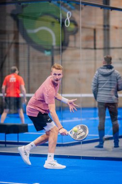 Riga, Letonya - 5 Nisan 2025 - Pembe tişörtlü odaklı padel oyuncusu, bir padel turnuvası sırasında kapalı mavi kortta, cam duvarın yanında düşük bir topu geri getiriyor..