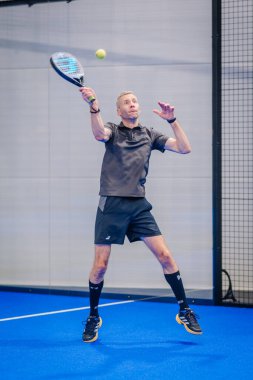 Riga, Letonya - 5 Nisan 2025 - Siyah kıyafetli Padel oyuncusu kapalı bir padel turnuvası maçında topu ezmeye hazırlanıyor..