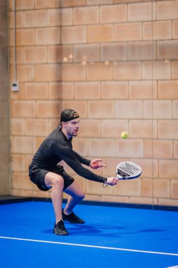 Riga, Letonya - 5 Nisan 2025 - Siyah elbiseli ve şapkalı erkek Padel turnuvası sırasında açık mavi bir iç sahada düşük bir atış yapmaya hazırlanıyor..