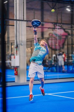 Riga, Letonya - 5 Nisan 2025 - Mavi tişörtlü erkek padel oyuncusu yoğun bir padel turnuvası sırasında kapalı alanda topu kırmak için zıplıyor.