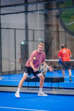 Riga, Letonya - 5 Nisan 2025 - Pembe tişörtlü bir erkek padel oyuncusu mavi bir iç sahada hızlı bir topu iade etmeye hazırlanıyor..