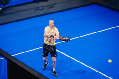 Riga, Letonya - 5 Nisan 2025 - Bej tişörtlü erkek padel oyuncusu, kapalı alanda oynanan bir turnuva sırasında mavi bir forehand vuruşu yaptı.