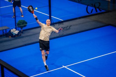 Riga, Letonya - 5 Nisan 2025 - Padel oyuncusu kapalı sahadaki bir turnuva sırasında topa hassas bir şekilde vurmak için havada zıplar..