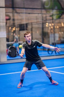 Riga, Letonya - 5 Nisan 2025 - Odaklanmış erkek padel oyuncusu canlı bir mavi kort turnuvasında kapalı alanda oynanan bir maç sırasında tekrar atış yapmaya hazırlanıyor..