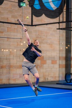 Riga, Letonya - 5 Nisan 2025 - Kapalı alan padel turnuvası sırasında güçlü bir vuruş yaparak enerji ve atletizm gösterisi yapan erkek padel oyuncusu.