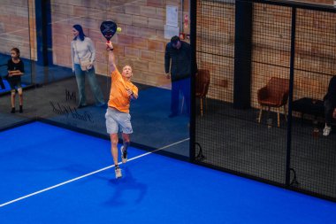 Riga, Letonya - 5 Nisan 2025 - Turuncu tişörtlü erkek padel oyuncusu, kapalı bir padel turnuvası sırasında yüksek topa vurmak için atlar ve seyirciler cam duvardan izlerler..