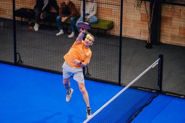 Riga, Letonya - 5 Nisan 2025 - Turuncu tişörtlü erkek padel oyuncusu yoğun kapalı alan padel turnuvası sırasında topu kırmak için ağdan atlıyor, arka planda seyirciler bulanıklaşıyor.