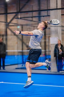 Riga, Letonya - 5 Nisan 2025 - Erkek Padel oyuncusu kapalı alanda oynanan bir turnuva maçında topu çarpmak için yüksek atlıyor..