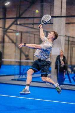 Riga, Letonya - 5 Nisan 2025 - Erkek Padel oyuncusu kapalı alanda oynanan bir turnuva maçında topu çarpmak için yüksek atlıyor..
