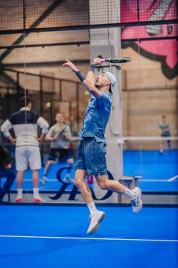 Riga, Letonya - 5 Nisan 2025 - Erkek padel oyuncusu bir padel turnuvasında atlamak için yükseğe sıçradı, çeviklik ve güçlü atletik performans sergiledi..