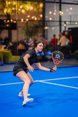 Riga, Letonya - 5 Nisan 2025 - Kadın Padel oyuncusu, canlı mavi bir iç sahada düzenlenen Padel turnuvasında sert bir vuruş yapmaya hazırlanıyor..