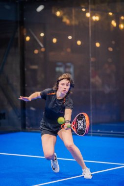 Riga, Letonya - 5 Nisan 2025 - Odaklanmış kadın padel oyuncusu, kapalı mavi bir sahada yapılan bir turnuva sırasında düşük vuruş yapmak için öne atılır..