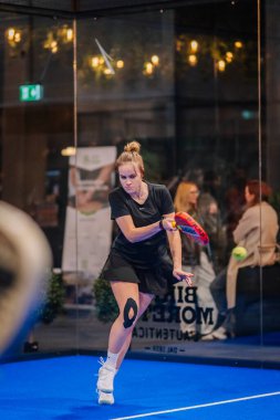 Riga, Letonya - 5 Nisan 2025 - Siyah kıyafetli kadın padel oyuncusu, kapalı alanda mavi bir kortta yakalanan Padel turnuvası sırasında düşük yoğunluklu bir atış yaptı..