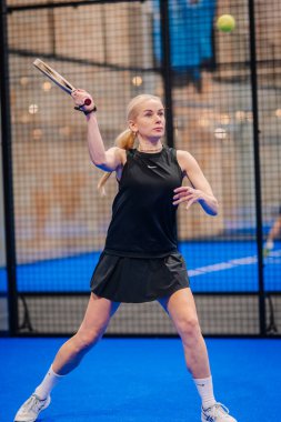 Riga, Letonya - 5 Nisan 2025 - Padel turnuvası sırasında kadın Padel oyuncuları, mavi iç sahada siyah spor kıyafetleri giyerek topa odaklanarak vurmaya hazırlanıyor..