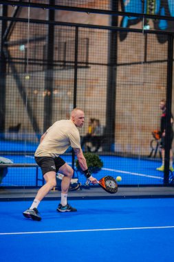 Riga, Letonya - 5 Nisan 2025 - Padel oyuncusu, mavi iç sahada oynanan, krema ve siyah spor kıyafetleri giymiş bir turnuva maçında düşük şiddette topa vuruyor..