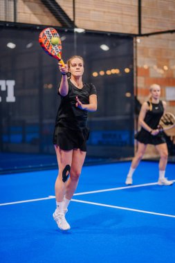 Riga, Letonya - 5 Nisan 2025 - Padel turnuvası sırasında bayan Padel oyuncusu topa odaklanmış bir şekilde vuruyor, takım arkadaşı da kapalı alanda mavi bir kortta izliyor..
