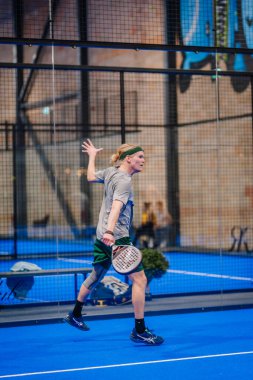 Riga, Letonya - 5 Nisan 2025 - Padel turnuvası sırasında elinde kendinden emin vücut diliyle raketini tutarak iç sahada yürüyen erkek padel oyuncusu.