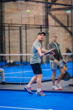Riga, Letonya - 5 Nisan 2025 - Padel turnuvası sırasında bir başka oyuncu kapalı sahadaki cam duvarın yanında izlerken erkek Padel oyuncusunun ön atış hakkı vardır..