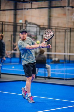 Riga, Letonya - 5 Nisan 2025 - Padel turnuvası sırasında bir başka oyuncu kapalı sahadaki cam duvarın yanında izlerken erkek Padel oyuncusunun ön atış hakkı vardır..