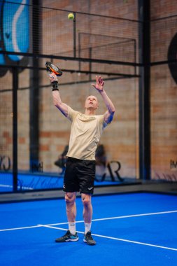 Riga, Letonya - 5 Nisan 2025 - Erkek Padel oyuncusu bir padel turnuvası sırasında servis yapmaya hazırlanıyor, topu mavi bir iç sahada krema ve siyah dişli bir şekilde fırlatıyor..