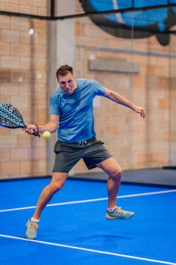 Riga, Letonya - 5 Nisan 2025 - Erkek padel oyuncusu kapalı alanda oynanan bir turnuva maçında topa vuruyor, yoğun ve orta hava atletik hareketlerini gösteriyor.