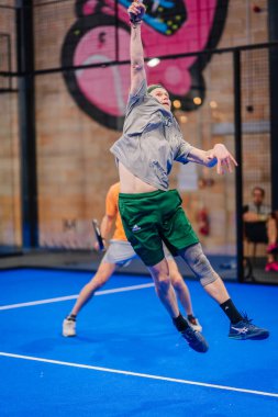 Riga, Letonya - 5 Nisan 2025 - Padel turnuvası sırasında erkek padel oyuncusu, kapalı mavi bir kortta yeşil şort ve gri gömlek giyerek yüksek vuruş yapmak için atlıyor..