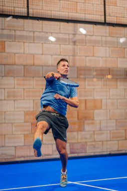 Riga, Letonya - 5 Nisan 2025 - Orta sahadaki erkek padel oyuncusu kapalı sahadaki bir turnuvada güçlü bir forehand vuruyor, atletizm ve odaklanma gösteriyor.
