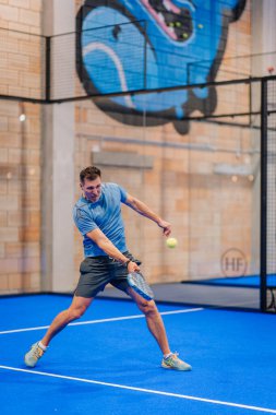 Riga, Letonya - 5 Nisan 2025 - Erkek padel oyuncusu kapalı alanda oynanan bir turnuva maçında topa vuruyor, yoğun ve orta hava atletik hareketlerini gösteriyor.