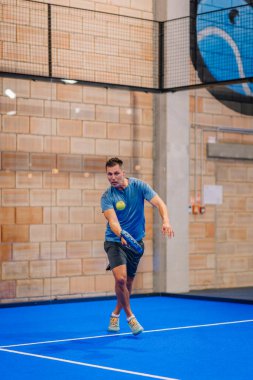 Riga, Letonya - 5 Nisan 2025 - Padel oyuncusu kapalı mavi bir sahada mavi tişört ve siyah şort giyen bir Padel turnuvası sırasında odaklanmış ters vuruş yaptı..