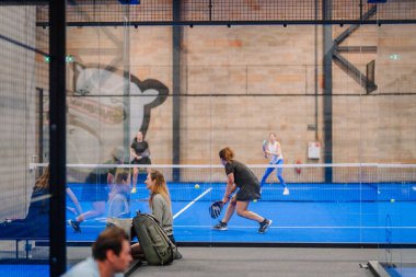 Riga, Letonya - 5 Nisan 2025 - Padel turnuvası sırasında iç sahada kadın padel oyuncuları eylem halindeyken, diğerleri cam muhafazanın yanında durup dinlenirken,.