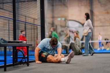 Riga, Letonya - 5 Nisan 2025 - Padel oyuncusu, diğer katılımcılar ve izleyiciler tarafından çevrelenen bir Padel turnuvası sırasında rahatsız ve muhtemelen yaralı bir şekilde yerde oturuyor..