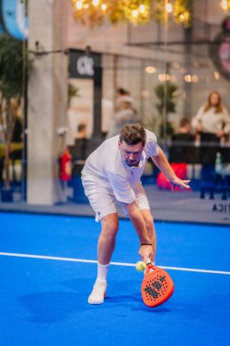 Riga, Letonya - 5 Nisan 2025 - Beyaz elbiseli erkek padel oyuncusu, kapalı alandaki bir sahada düzenlenen bir Padel turnuvası sırasında düşük bir topa vururken yoğun bir şekilde odaklanır..