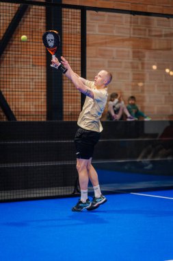 Riga, Letonya - 5 Nisan 2025 - Padel oyuncusu atletizm formunu gösteren ve kapalı alanda oynanan mavi sahaya odaklanan yüksek bir topa vurmak için atlar..