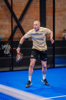 Riga, Letonya - 5 Nisan 2025 - Padel oyuncusu, mavi iç sahada oynanan bir turnuva sırasında krem rengi gömlek ve odaklı ifade ile siyah şort giyerek atış yapmaya hazırlanıyor..