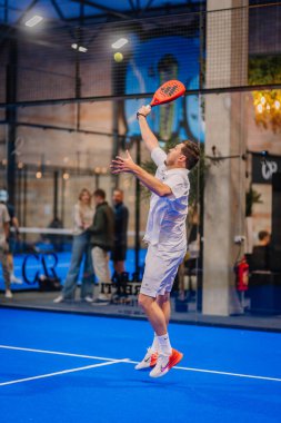 Riga, Letonya - 5 Nisan 2025 - Padel turnuvası sırasında beyaz elbise ve turuncu ayakkabılar giyen erkek padel oyuncusu, kapalı alanda mavi bir kortta atlamak için atlıyor..