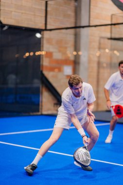 Riga, Letonya - 5 Nisan 2025 - Beyaz elbiseli erkek padel oyuncusu, bir padel turnuvası maçında düşük topa vurmak için esniyor, çevikliğini gösteriyor ve mavi iç saha sahasına odaklanıyor..