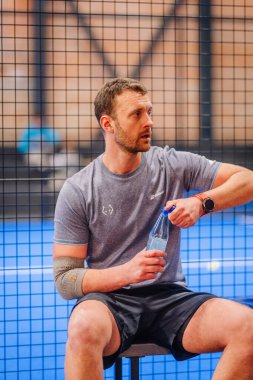 Riga, Letonya - 5 Nisan 2025 - Padel turnuvası sırasında erkek padel oyuncusu saha kenarında oturur, maçlar arasında dinlenirken su şişesi açar, spor kıyafetleri giyer ve dirsek desteği takar..