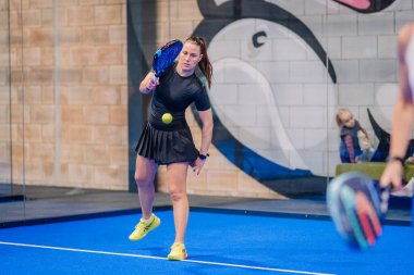 Riga, Letonya - 5 Nisan 2025 - Padel turnuvası sırasında top havada ve arka planda seyirciler varken forehand vuruşu yapan kadın padel oyuncusu.