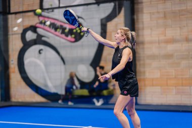 Riga, Letonya - 5 Nisan 2025 - Siyah kıyafetli odaklanmış kadın padel oyuncusu arka planda duvar resmiyle kapalı bir padel turnuvası sırasında üstten çekim yapıyor..