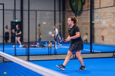 Riga, Letonya - 5 Nisan 2025 - Padel oyuncusu kapalı alanda yapılan bir turnuva sırasında net arkasında canlı bir mavi kort üzerinde durarak dönüş vuruşuna odaklanır..