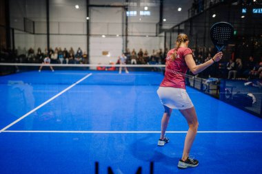 Riga, Letonya - 5 Nisan 2025 - Padel 'de yarışan bir Padel turnuvası sırasında bir bayan oyuncu, maçı camların arkasından izleyen bir kalabalıkla servis yapmaya hazırlanıyor..