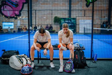 Riga, Letonya - 5 Nisan 2025 - İki bayan Padel oyuncusu, yanlarında ekipman ve çantalarla kibrit, su ve iyileşme arasında bir bankta dinleniyor..