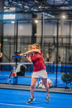 Riga, Letonya - 5 Nisan 2025 - Kırmızı tişörtlü ve beyaz etekli odaklanmış kadın padel oyuncusu, kapalı bir sahada düzenlenen yoğun bir padel turnuvası sırasında güçlü bir forehand vuruyor..
