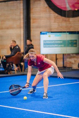 Riga, Letonya - 5 Nisan 2025 - Padel oyuncusu, arka planda skorbordlar ve izleyiciler ile rekabetçi bir Padel turnuvası maçında topu geri vermek için alçaktan geriliyor.