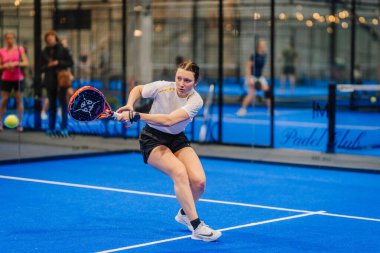 Riga, Letonya - 5 Nisan 2025 - Kadın Padel oyuncusu, kapalı alanda oynanan bir turnuva maçında düşük topa doğru hamle yapıyor..